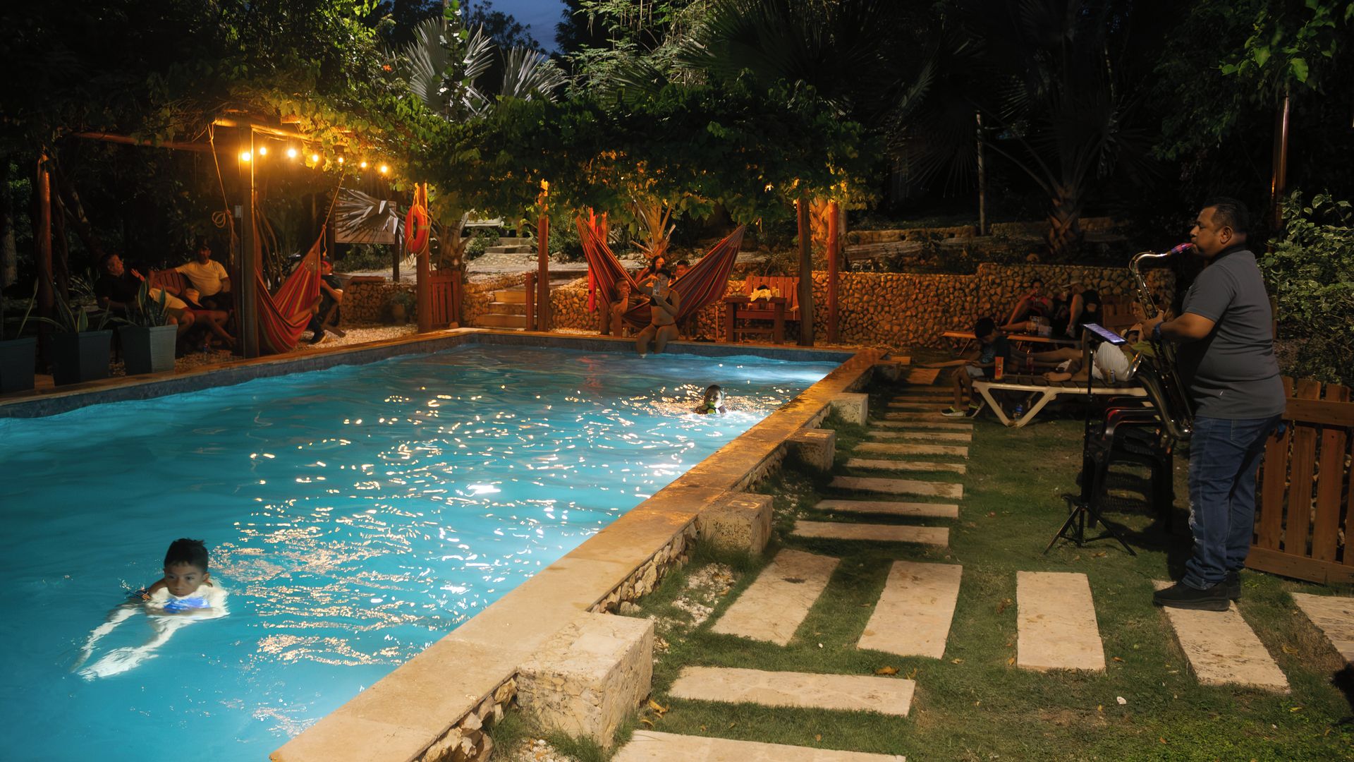A swimming pool with a stone border, surrounded by a stone wall and a grassy area with steps leading to a wooden deck.