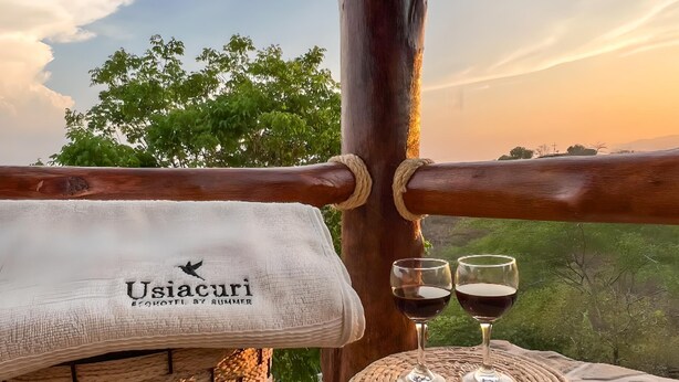 Una cesta de mimbre, una toalla con la marca 'Usiacuri' y dos copas de vino sobre un alféizar de piedra con vistas a árboles y al atardecer.