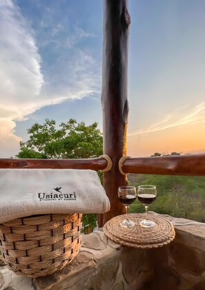 Un panier en osier, une serviette ornée du logo « Usiacuri », et deux verres à vin posés sur une corniche en pierre, d’où l’on peut admirer les arbres et le coucher de soleil.