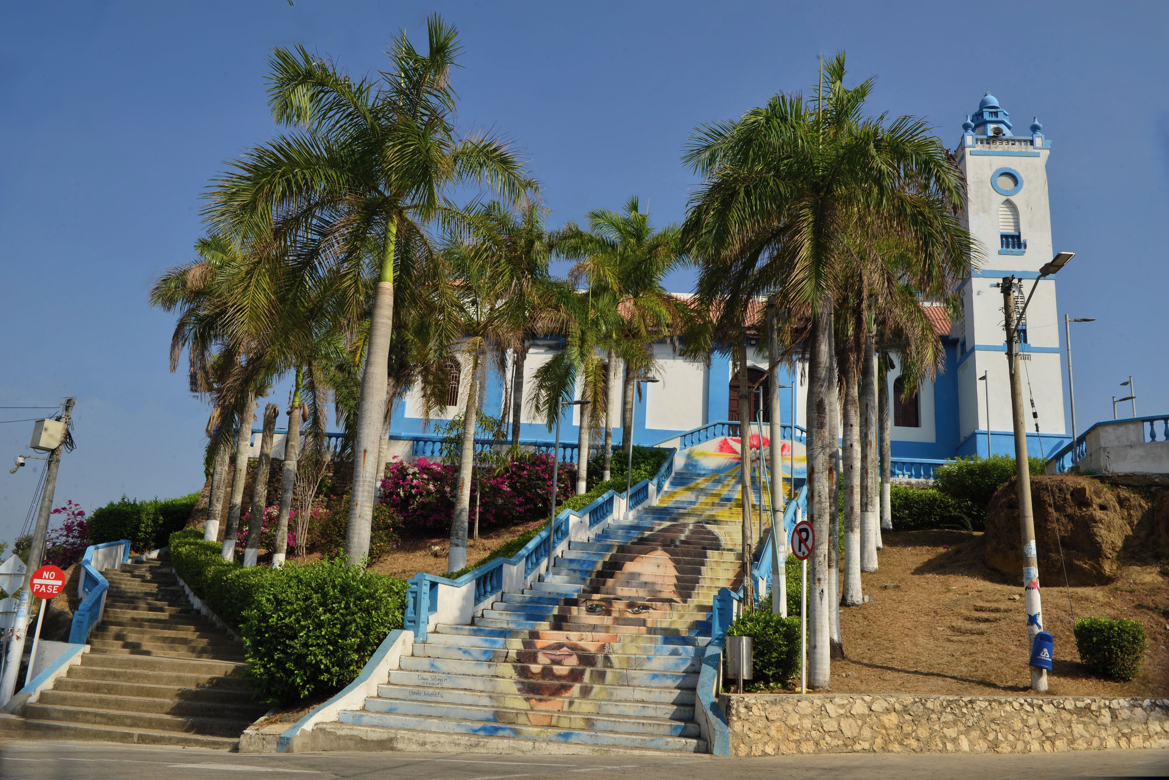 A staircase leading to a building with a clock tower, surrounded by palm trees and a clear sky.