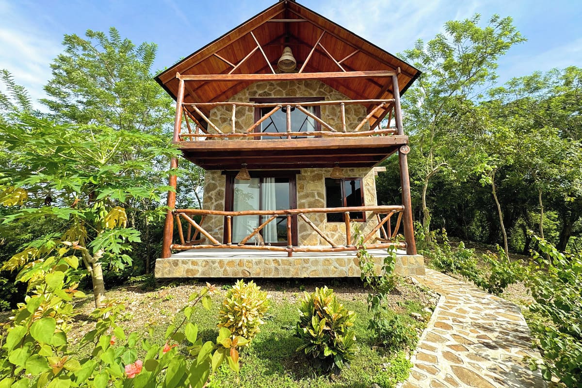 Une cabane en bois à deux étages, avec des fondations en pierre, entourée d’une végétation luxuriante.