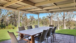 An outdoor covered patio with a wooden table and wicker chairs, surrounded by a lush garden and trees. - MarMel - Four bedroom home with pool and fire pit (Kangaroo Valley)
