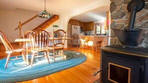 A cozy living room with a wooden dining table, chairs, and a blue patterned rug. A stone fireplace with a black stove is visible. - Newfound Lake Hideaway Mooring included (Bristol)