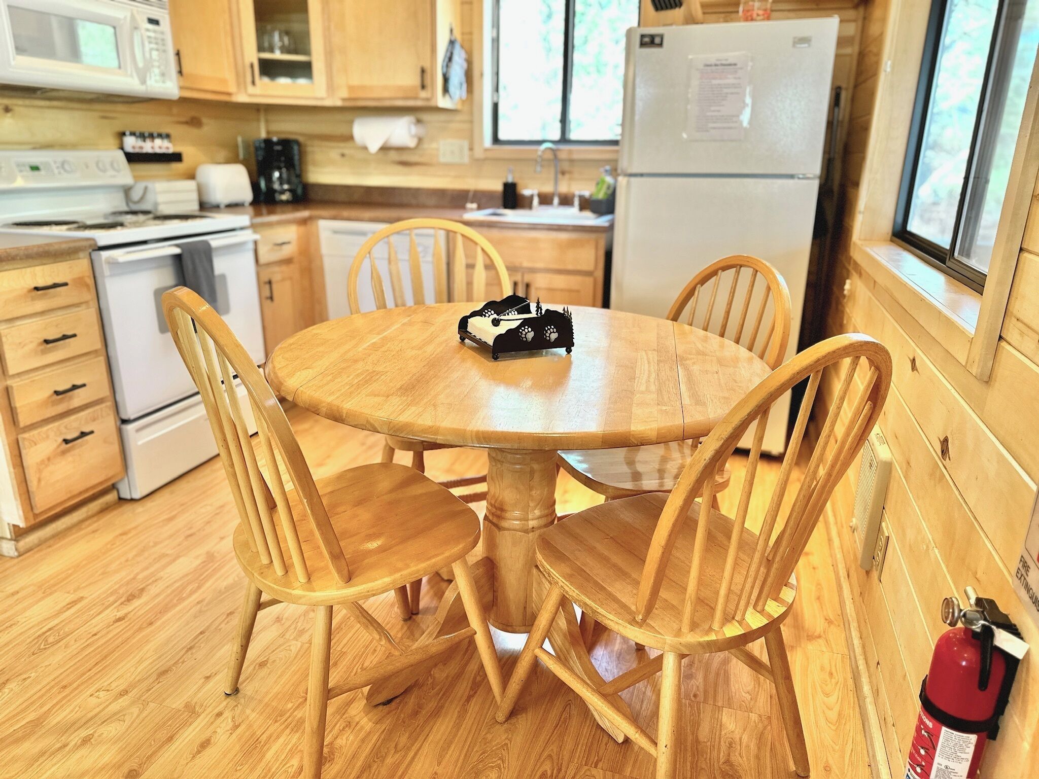 Une table de salle à manger en bois, accompagnée de quatre chaises, se trouve dans une cuisine dotée de meubles en bois, d’un réfrigérateur et d’une fenêtre.