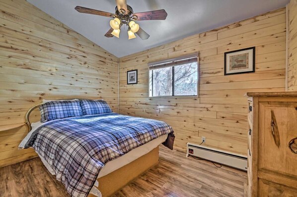 Een slaapkamer met houten wanden, een bed met geruit beddengoed, een plafondventilator, een raam en een houten dressoir.