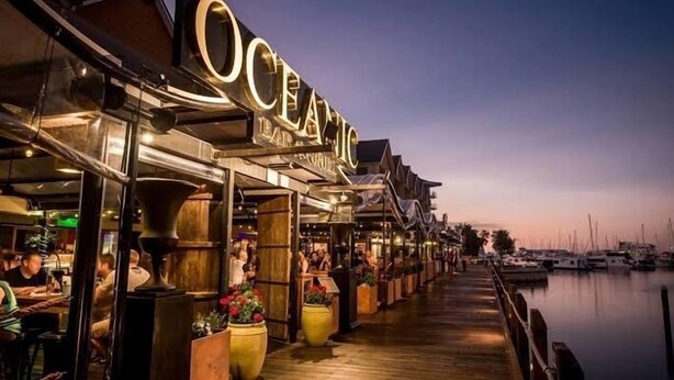 Un restaurant situé au bord de l’eau, doté d’une terrasse en bois et de tables en plein air, offrant une vue sur un port de plaisance rempli de bateaux.
