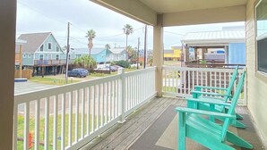 A covered porch with a view of a residential street, wooden deck, and turquoise chairs.