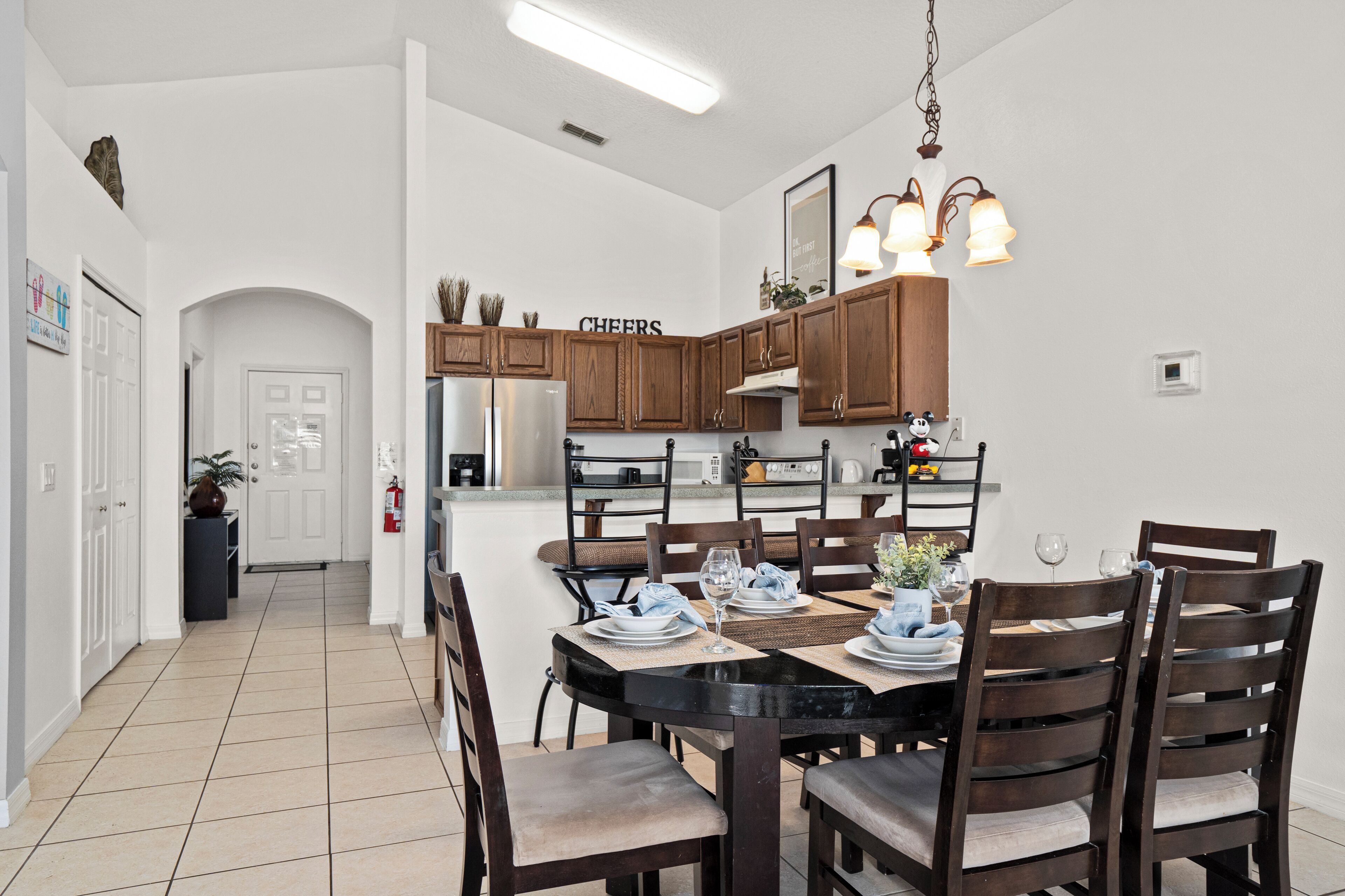 A modern kitchen with wooden cabinets, a dining table set for six, and a 'CHEERS' sign above the cabinets.