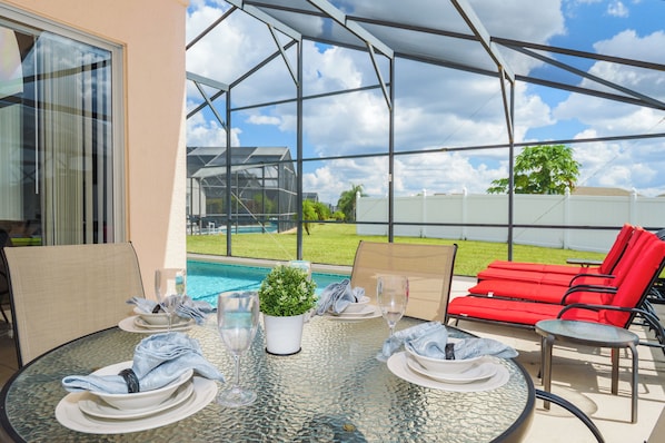 A covered outdoor dining area with a glass table, red lounge chairs, and a view of a swimming pool and greenery. - Beautiful, sun-drenched 4 Bed 3 Bath Villa within The Sanctuary at West Haven (Davenport)