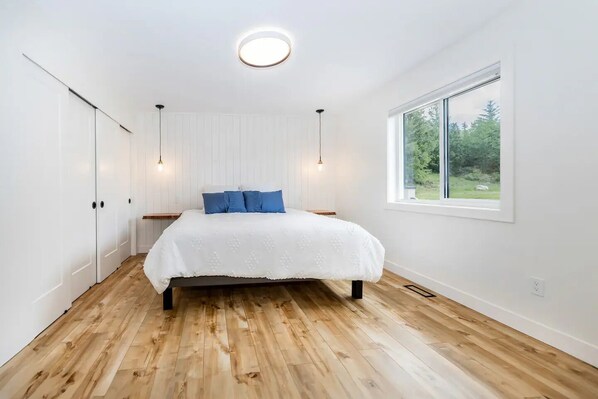 A bedroom with a wooden floor, a bed with a white bedspread and blue pillows, a window with a view of greenery, and a ceiling light. - Chalet Firefly BC REGISTRY APPROVAL #H631676206 (Halfmoon Bay)
