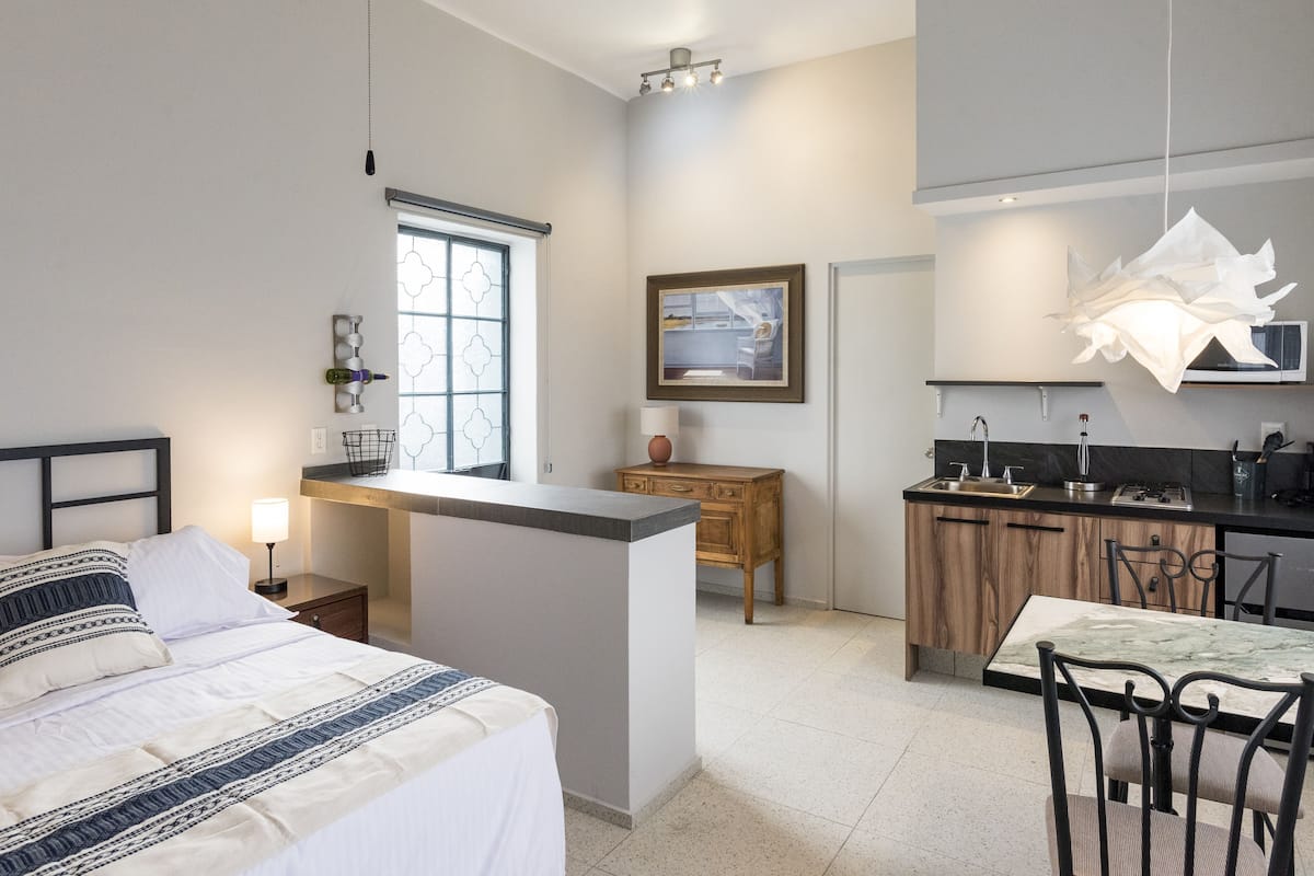 A modern hotel room with a bed, bedside table, lamp, and a view of the kitchen area.
