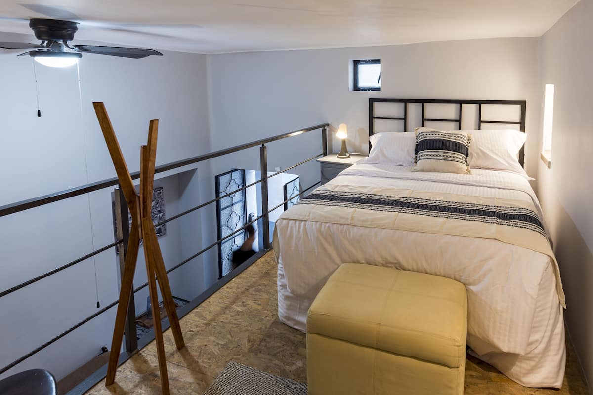 A bedroom with a bed, bedside table, lamp, and a view of the outside.