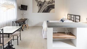 A compact hotel room with a bed, a desk with a chair, a TV, and a large window with curtains.