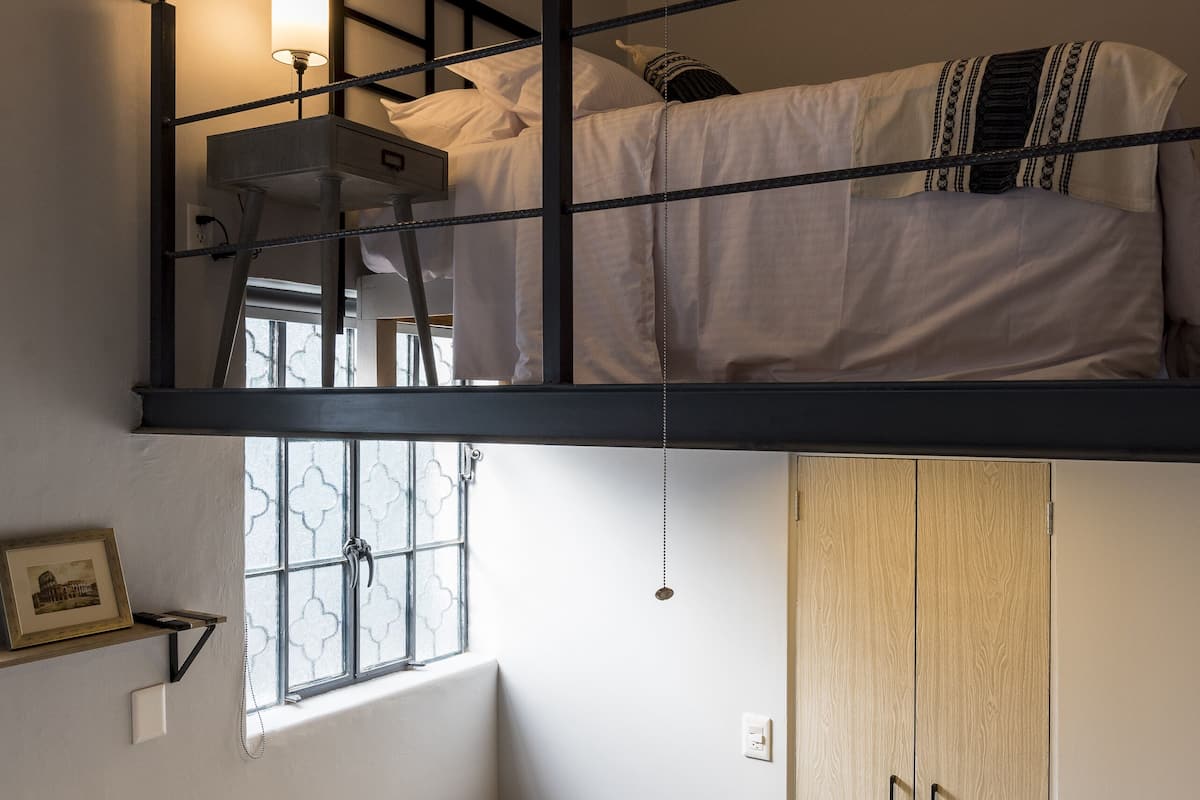 A loft-style bedroom with a bunk bed, a desk with a lamp, a chair, a small table, and a wardrobe.