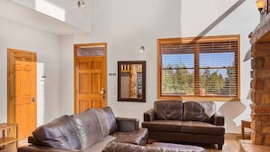A spacious living room with leather sofas, a wooden door, a large window with blinds, and a stone fireplace. - Large Log Home Near Zion (Orderville)