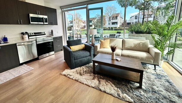 A modern kitchen with dark cabinets, a microwave, and a stove. There is a black armchair with a yellow pillow and a beige sofa with a decorative vase. A wooden coffee table is in front of the sofa. The room has large windows with a view of a residential area. - Clean & stylish with exclusive patio, king bed, secured parking, by the beach  (San Diego)