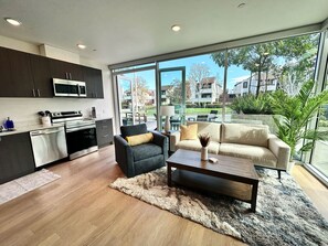 A modern kitchen with dark cabinets, a microwave, and a stove. There is a black armchair with a yellow pillow and a beige sofa with a decorative vase. A wooden coffee table is in front of the sofa. The room has large windows with a view of a residential area.