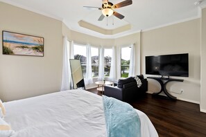 A hotel room with a large bed, a ceiling fan, a TV, a sofa, and a painting of a beach scene.