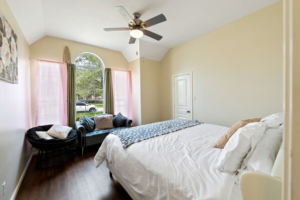A bedroom with a bed, a window with curtains, a ceiling fan, and a view of the outside.