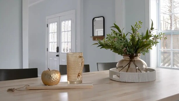 Et spiseområde med et træbord, en vase med grønne planter, et gyldent æble og en høj glasvase.