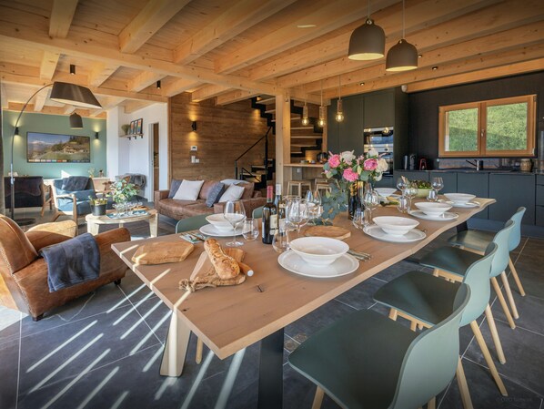 Amplia sala de estar con una mesa de comedor de madera dispuesta para una comida, rodeada de asientos cómodos y vistas a la cocina.