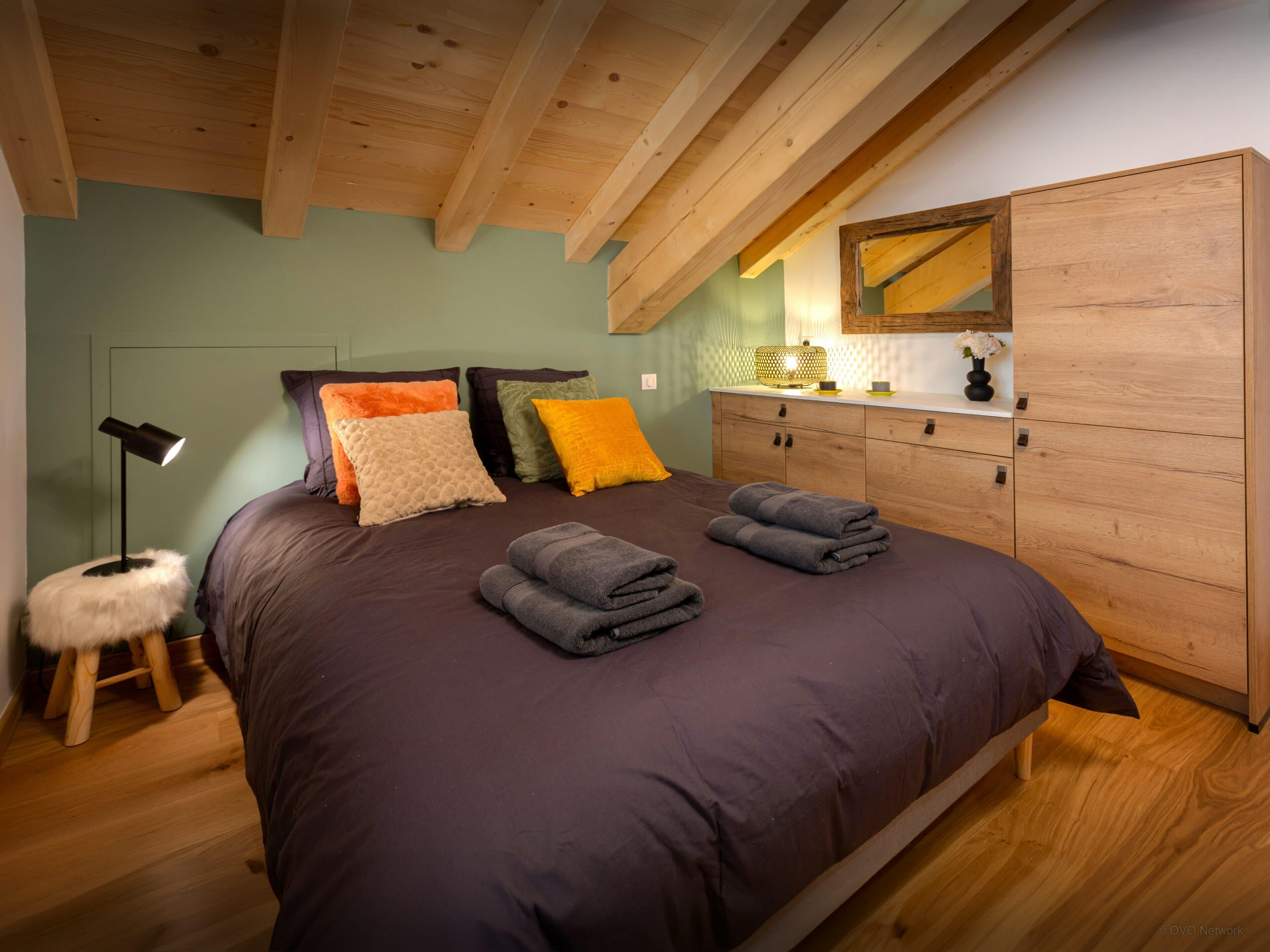 A bedroom with a wooden ceiling, a bed with multiple pillows, a bedside lamp, a small stool, and a wooden wardrobe.