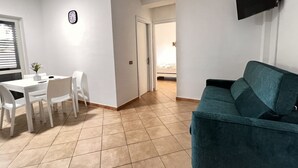 Ein Wohnzimmer mit einem türkisfarbenen Sofa, einem Essbereich mit weißem Tisch und Stühlen, einem an der Wand montierten Flachbildfernseher und einer Wanduhr.