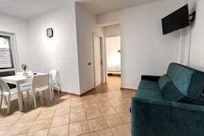 Un salon avec un canapé de couleur turquoise, un coin repas avec une table et des chaises blanches, un téléviseur à écran plat fixé au mur et une horloge au mur.