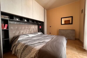 Ein Schlafzimmer mit einem gepolsterten Kopfteil, einem Bett mit gemusterten Betttextilien, einem Nachttisch, einer Kommode und einem Spiegel an der Wand.