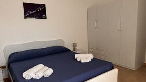 Ein ordentlich bezogenes Bett mit blauer Bettwäsche und weißen Handtüchern, eine Nachttischlampe und ein zweitüriger Kleiderschrank.