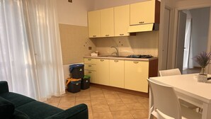 Eine kleine Küche mit gelben Schränken, einer weißen Arbeitsplatte und einem Spülbecken. Im Vordergrund steht ein Esstisch mit weißen Stühlen und ein grünes Sofa.