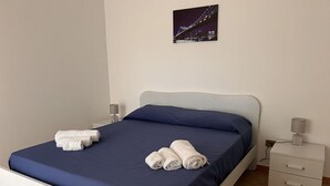 Un lit bien fait, avec des draps bleus et des serviettes blanches, une table de chevet avec une lampe et un tableau représentant un paysage urbain accroché au mur.