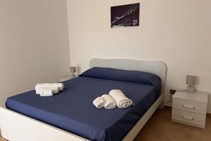 A neatly made bed with blue sheets and white towels, a nightstand with a lamp, and a cityscape picture on the wall.