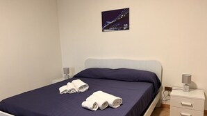 Ein ordentlich bezogenes Bett mit dunkelblauer Bettdecke und weißen Laken, ein Nachttisch mit Lampe und ein gerahmtes Bild einer Brücke an der Wand.