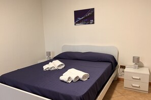 Ein ordentlich bezogenes Bett mit dunkelblauer Bettdecke und weißen Laken, ein Nachttisch mit Lampe und ein gerahmtes Bild einer Brücke an der Wand.