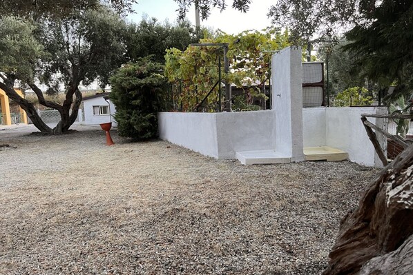 Un patio de grava con un muro blanco, un pequeño edificio blanco y un árbol.