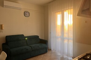 Una sala de estar con un sofá verde, un reloj en la pared y cortinas translúcidas en la ventana.