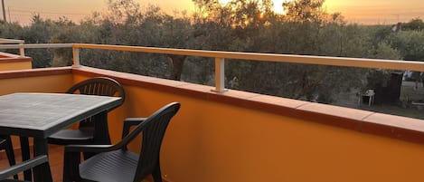 A balcony with a table and chairs, overlooking a landscape with trees and a sunset.