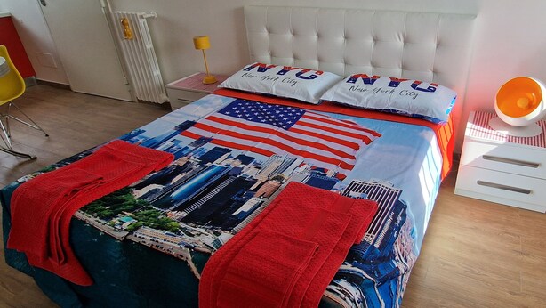 A hotel room with a bed featuring an American flag and 'NYC' pillows, a red blanket, a lamp, and a small white nightstand. - Special Guest Home (BARI)