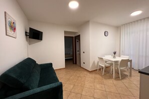 A living room with a green sofa, a TV mounted on the wall, a dining area with a table and chairs, and a kitchen area with a counter.