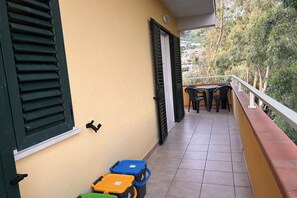 Un balcón con suelo de baldosas, una mesa con sillas y vistas a zonas verdes y edificios al fondo.