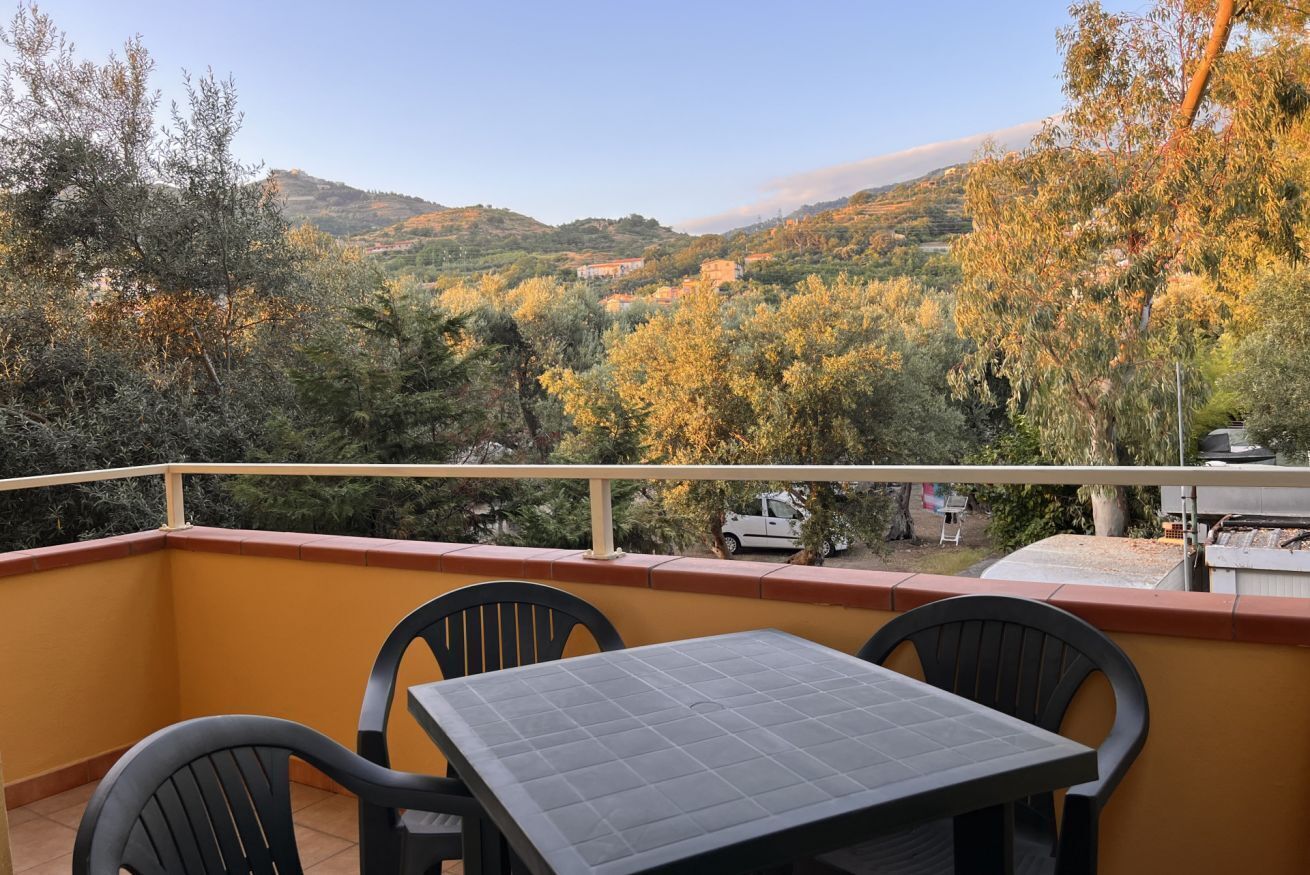Uma varanda com mesa e cadeiras, com vista para uma paisagem acidentada com árvores e edifícios.
