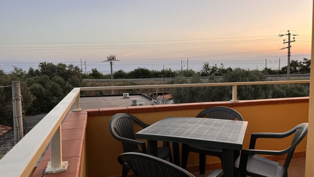 Ein Balkon mit Tisch und Stühlen, der auf eine Küstenlandschaft mit Vegetation und Stromleitungen blickt.