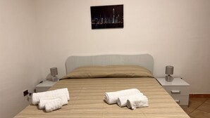 Ein ordentlich bezogenes Bett mit Streifenmuster, zwei weiße Handtücher, ein Nachttisch mit Lampe und ein Stadtbild an der Wand.