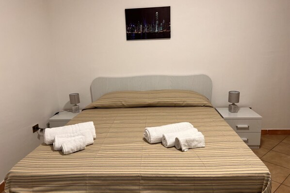 A neatly made bed with striped bedding, two white towels, a nightstand with a lamp, and a cityscape picture on the wall.