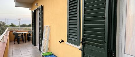 A balcony with a table and chairs, green shutters, and trash bins.