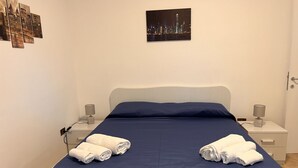 Un lit bien fait, recouvert d’une couvre-lit bleu foncé, avec deux serviettes blanches pliées au pied du lit. Il y a deux tables de chevet, chacune avec une lampe et un petit objet décoratif. Un tableau encadré représentant un paysage urbain est accroché au mur, au-dessus du lit.