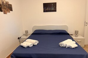 Een net opgemaakt bed met een donkerblauwe sprei en twee witte handdoeken gevouwen aan het voeteneinde. Twee nachtkastjes, elk met een lamp en een klein decoratief object. Een lijst met een stadsgezicht hangt aan de muur boven het bed.