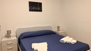 Ein ordentlich bezogenes Bett mit blauer Bettwäsche und weißen Handtüchern, ein Nachttisch mit einer Lampe und ein Stadtbild an der Wand.