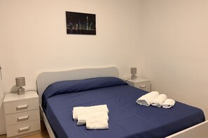 En pænt redt seng med blåt sengetøj og hvide håndklæder, et natbord med lampe, og et bybillede på væggen.